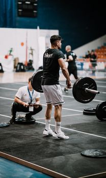 A muscular man lifting weights in a gym with a referee observing indoors, showcasing athletic strength.