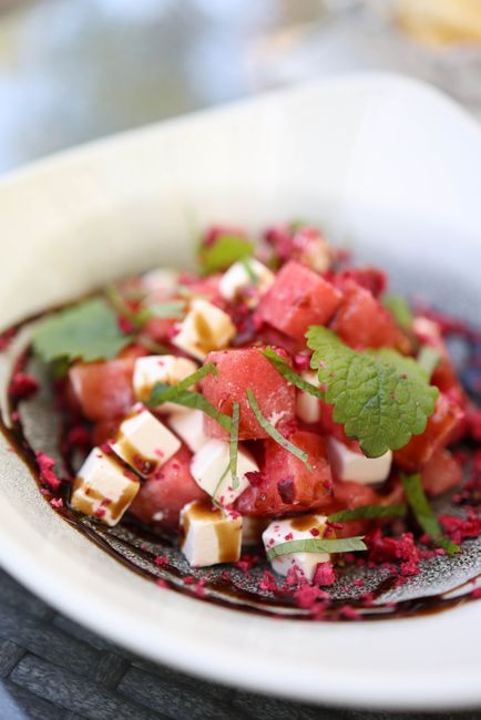 A wide white platter of cubed watermelon tossed with crumbled feta, thin red onion slices, and torn mint leaves, glistening with olive oil.