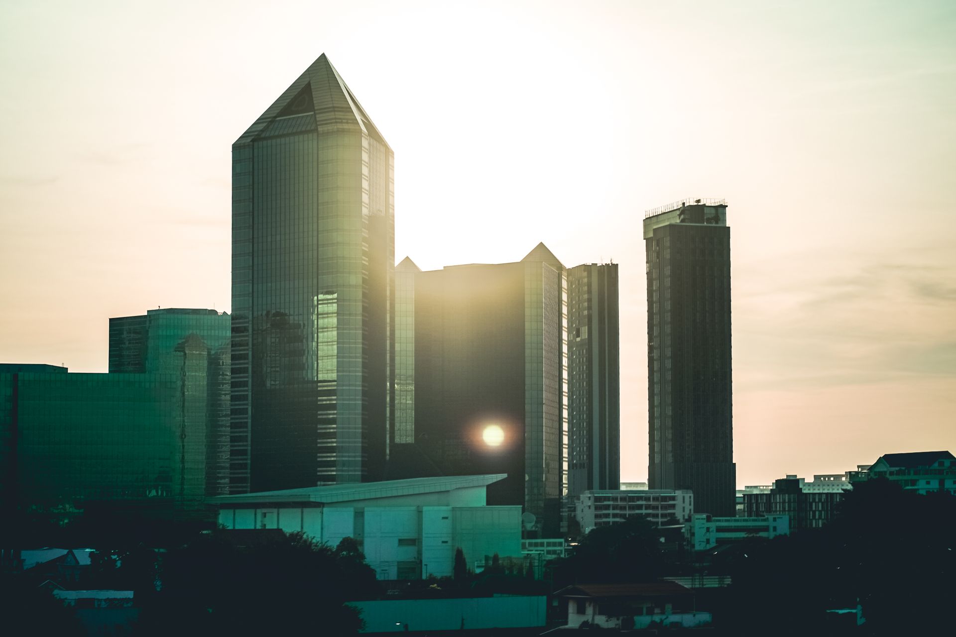Sunlight peeks through modern skyscrapers in Bangkok, Thailand, creating a dramatic urban skyline.
