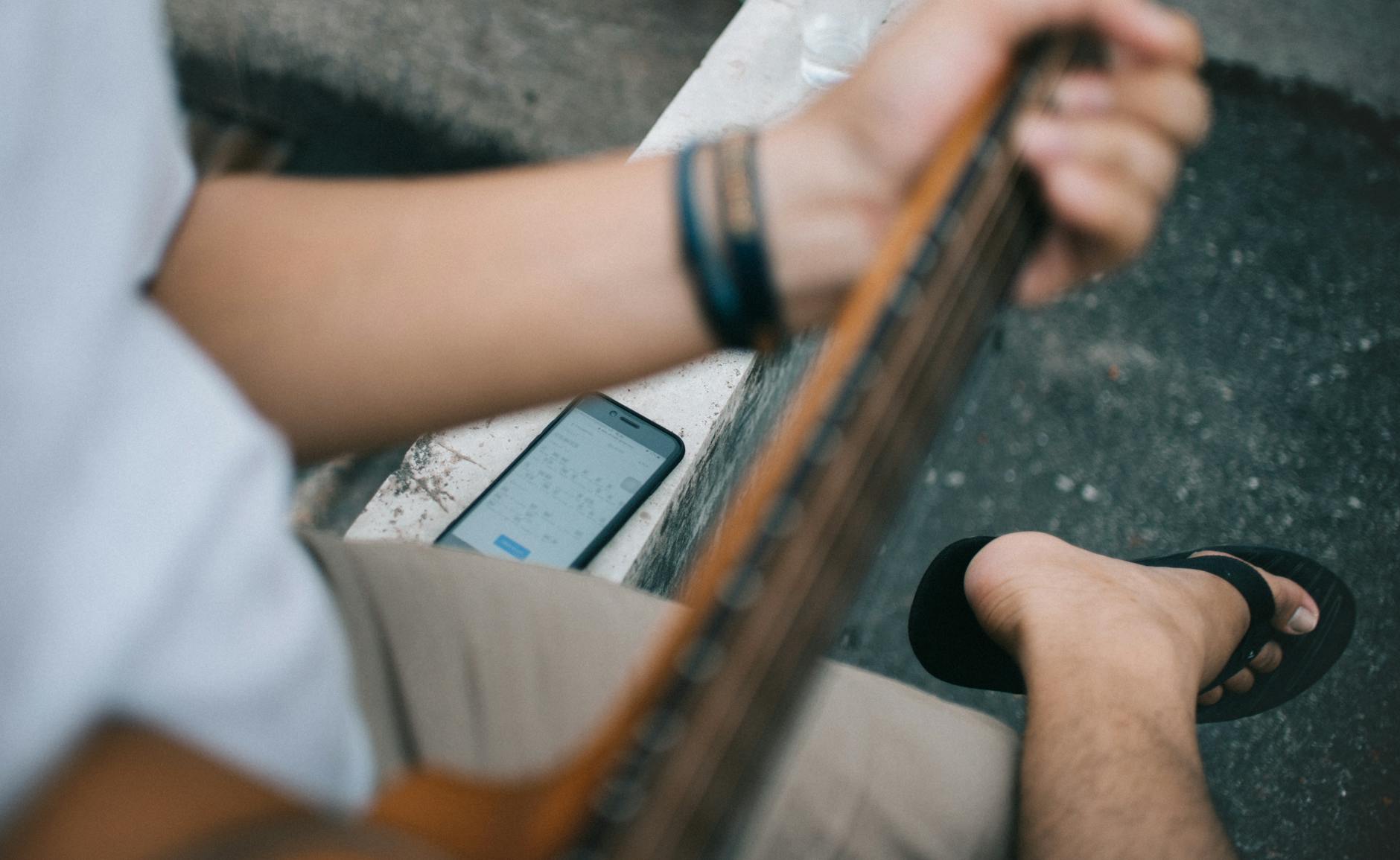 A young man plays a guitar outdoors, using his phone for notes. Casual and musical vibe.