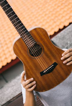 Close-up of a person holding a wooden acoustic guitar against a tiled rooftop background.
