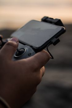 Detailed close-up of a hand holding a drone controller with a smartphone attached, outdoors at sunset.