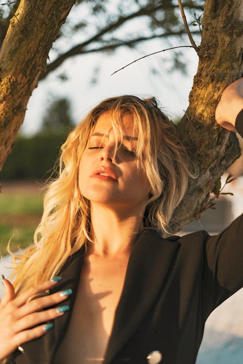 Young Woman in a Black Jacket Standing by a Tree in Direct Sunlight 