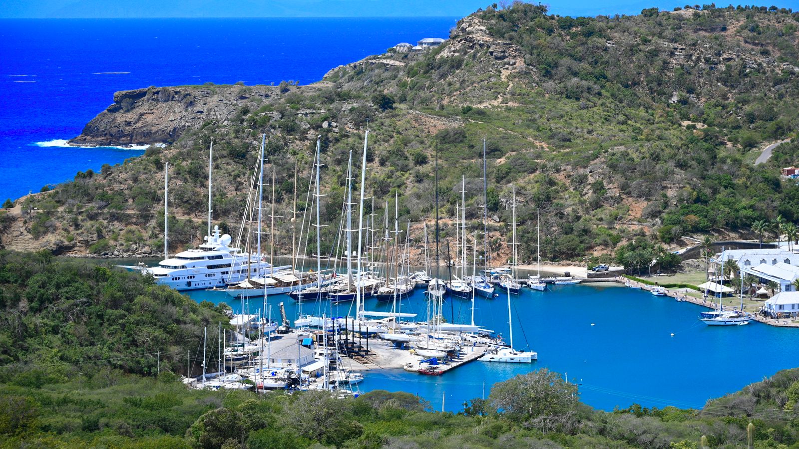 Stunning aerial view of a serene harbor with moored yachts, surrounded by lush hills and vibrant blue water.
