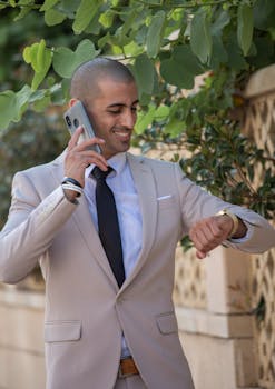 Professional businessman in a beige suit checks his watch while on a phone call outdoors.