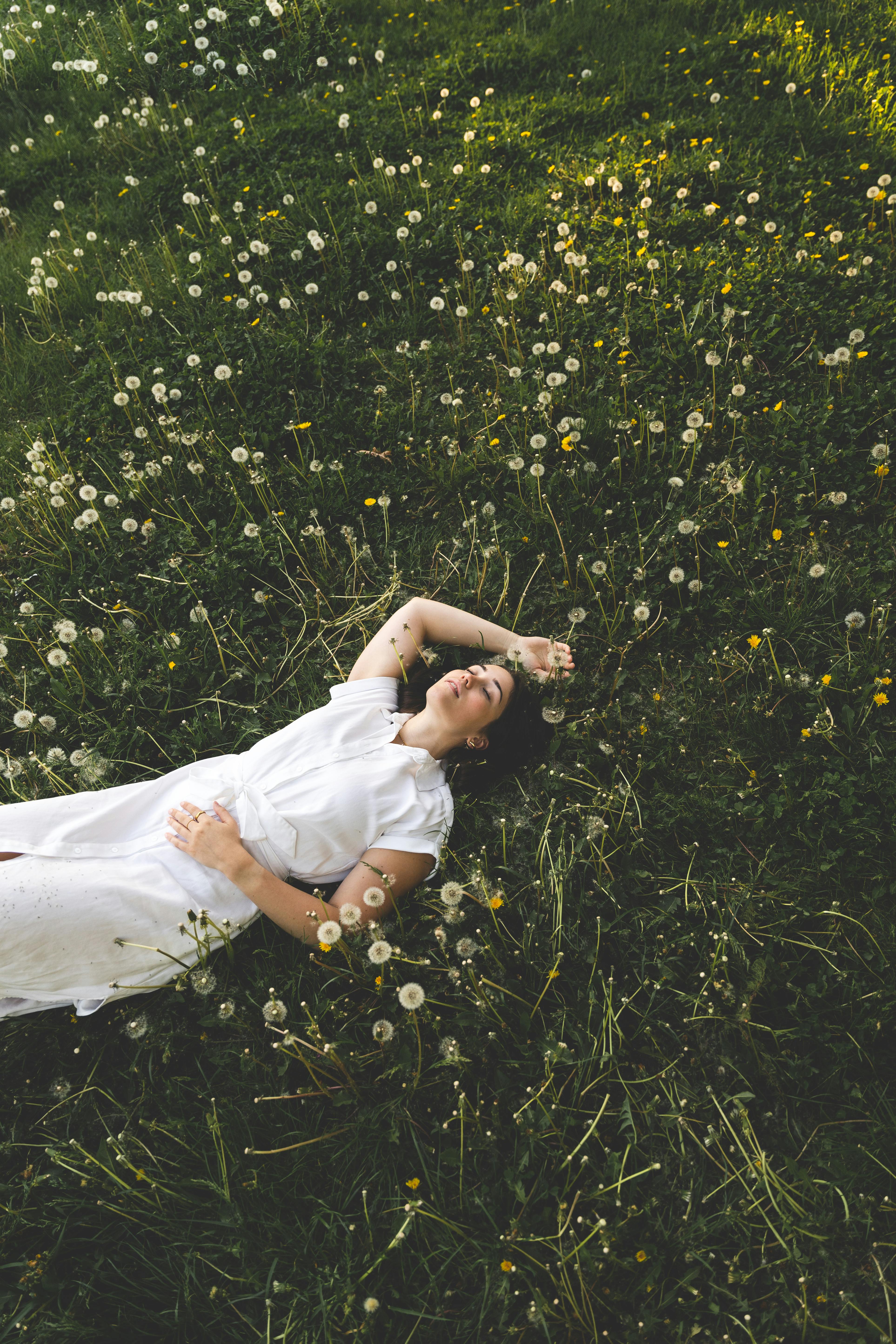 A woman in a white dress relaxes in a dandelion field at sunset, enjoying nature's tranquility.