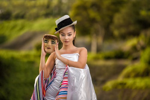 Young Woman in a Dress and Hat Standing Outside and Holding a Mask 