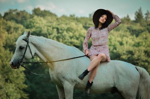 Free A woman in a stylish dress and hat sits elegantly on a white horse in a rural summer setting. Stock Photo