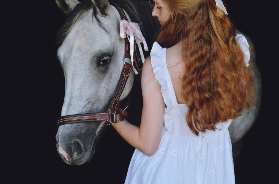 Redheaded woman in white dress with a horse against a dark backdrop.