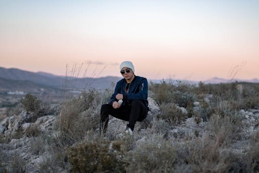 A fashionable man in a beanie and sunglasses sitting outdoors on rocky terrain during a pastel sunset.