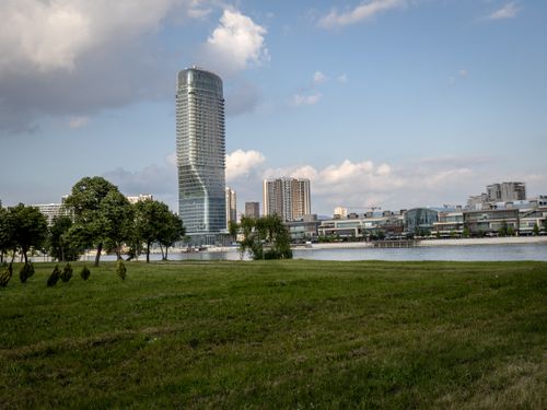 Free A modern skyscraper towers over a serene park with lush greenery and a calm lake. Stock Photo