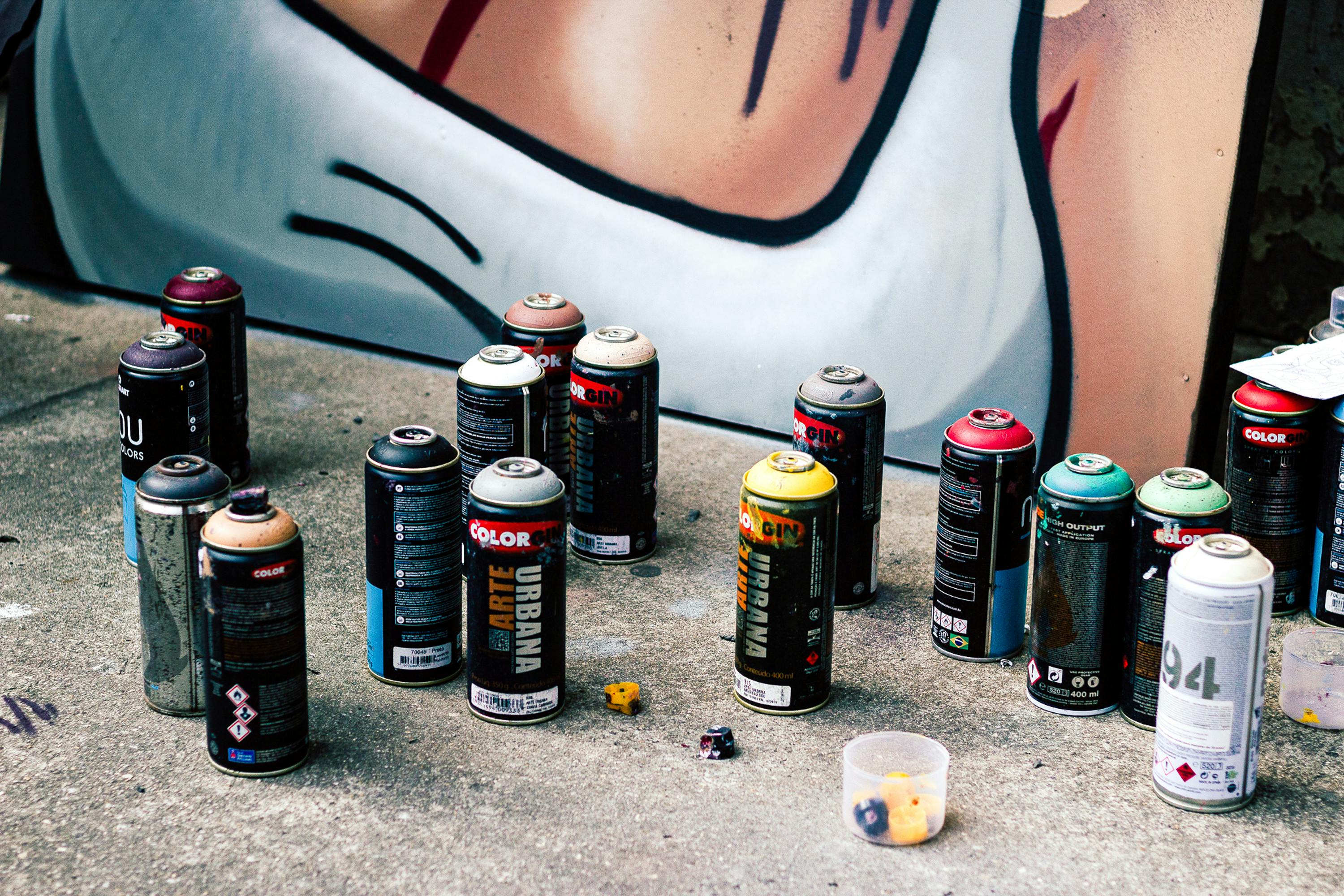 Colorful spray paint cans on concrete in front of a graffiti mural in São Paulo.