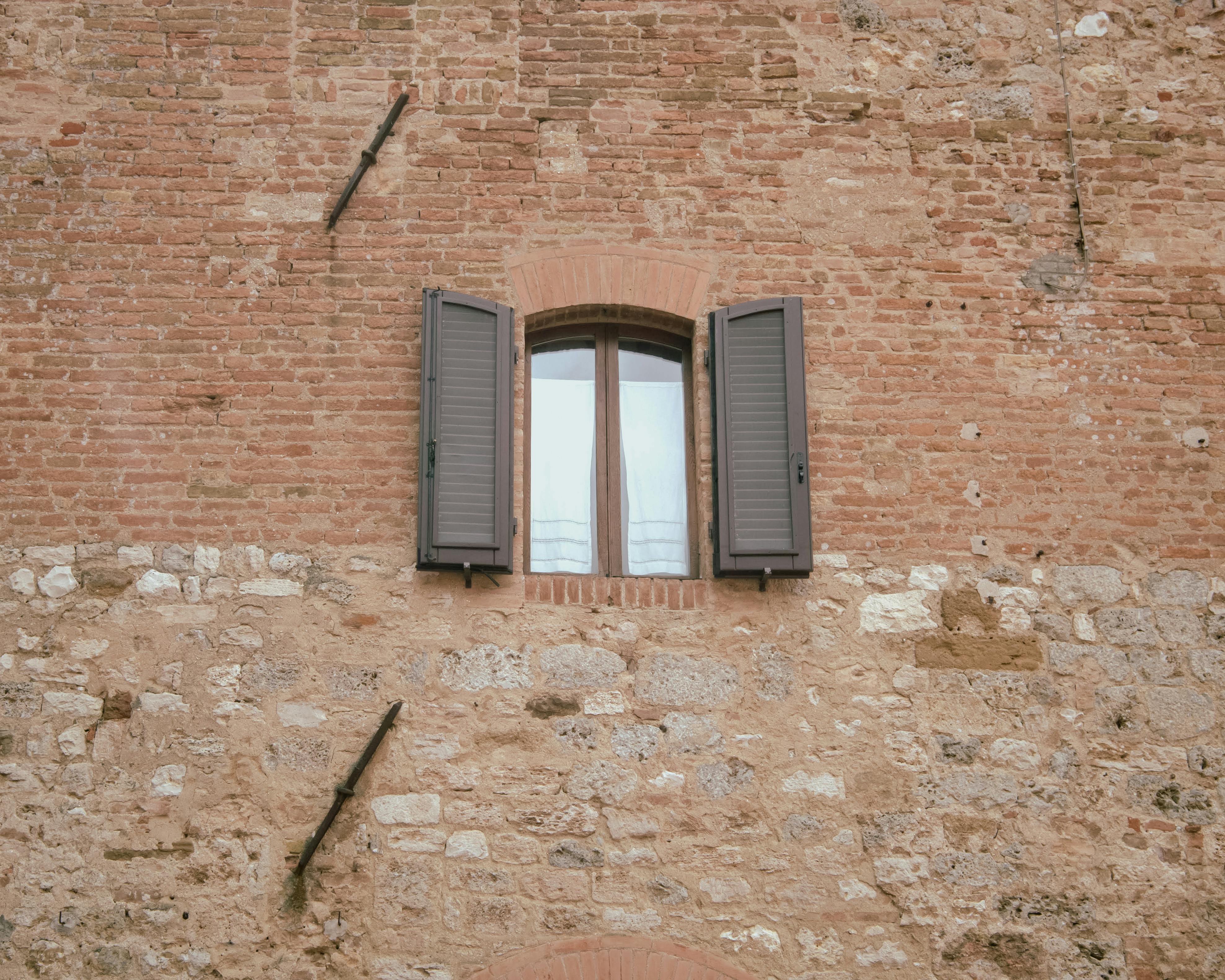 Vintage brick wall featuring an arched window with open wooden shutters, offering a glimpse of architectural charm.