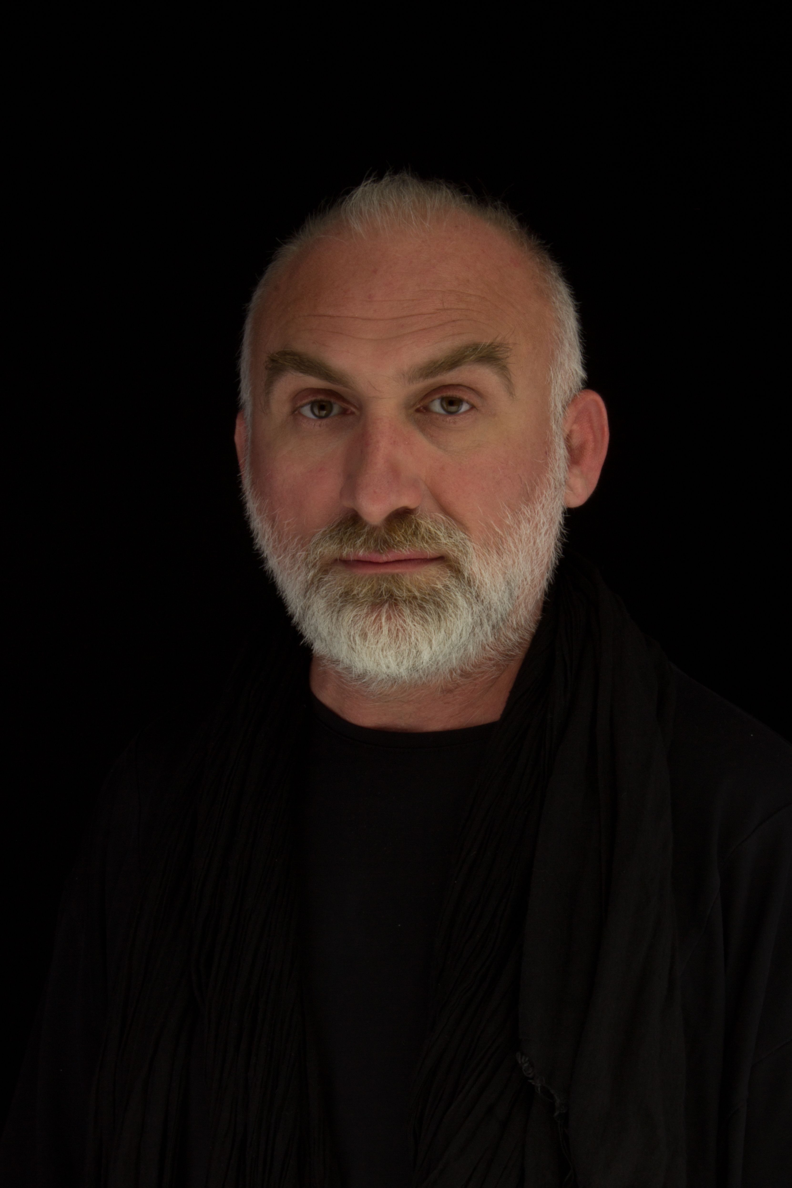 Striking studio portrait of an adult man with a white beard and mustache against a black background.