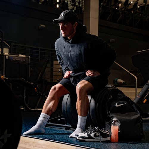 Muscular Man in a Hoodie and Cap Sitting at the Gym 