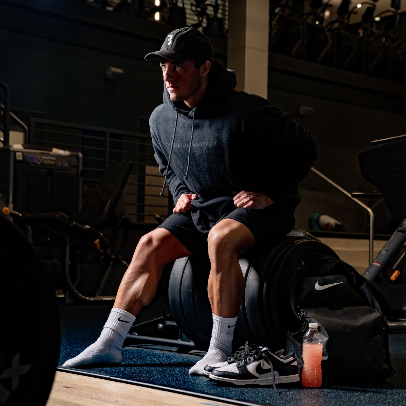 Hombre con hoodie y gorra descansando en gimnasio oscuro