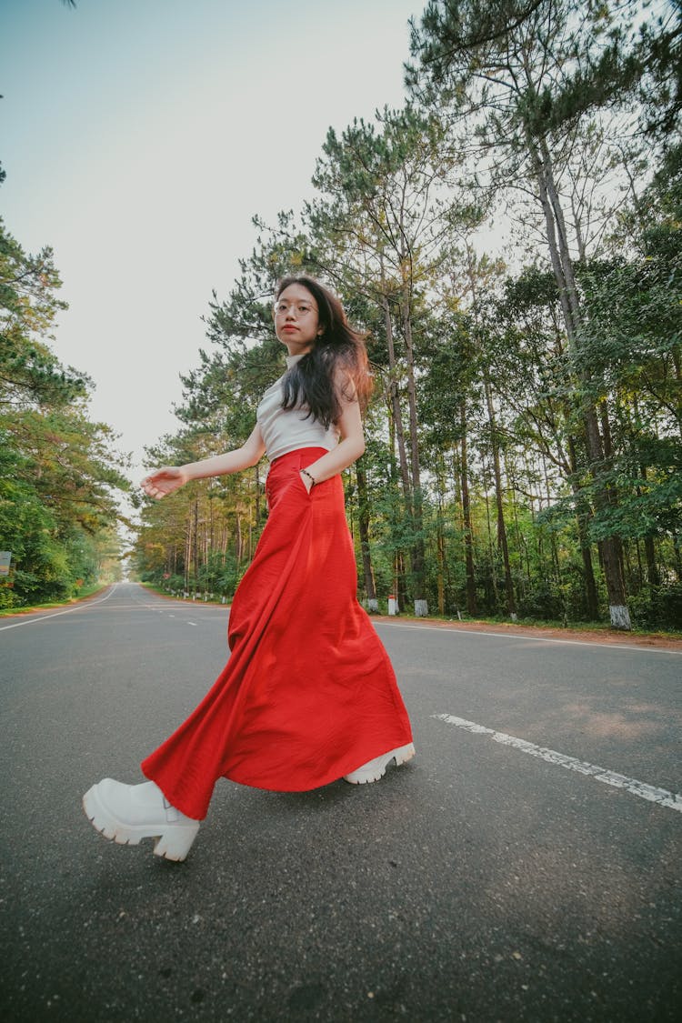 Young Woman In Long Satin Skirt Walking Across A Forest Road