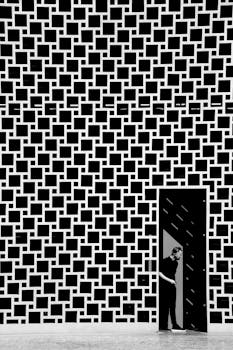 Black and white photo of a man exiting a modern geometric facade through a door.