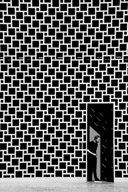Black and white photo of a man exiting a modern geometric facade through a door.