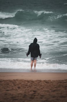 A person stands in the shoreline with waves approaching, wearing a hooded jacket during a windy day.