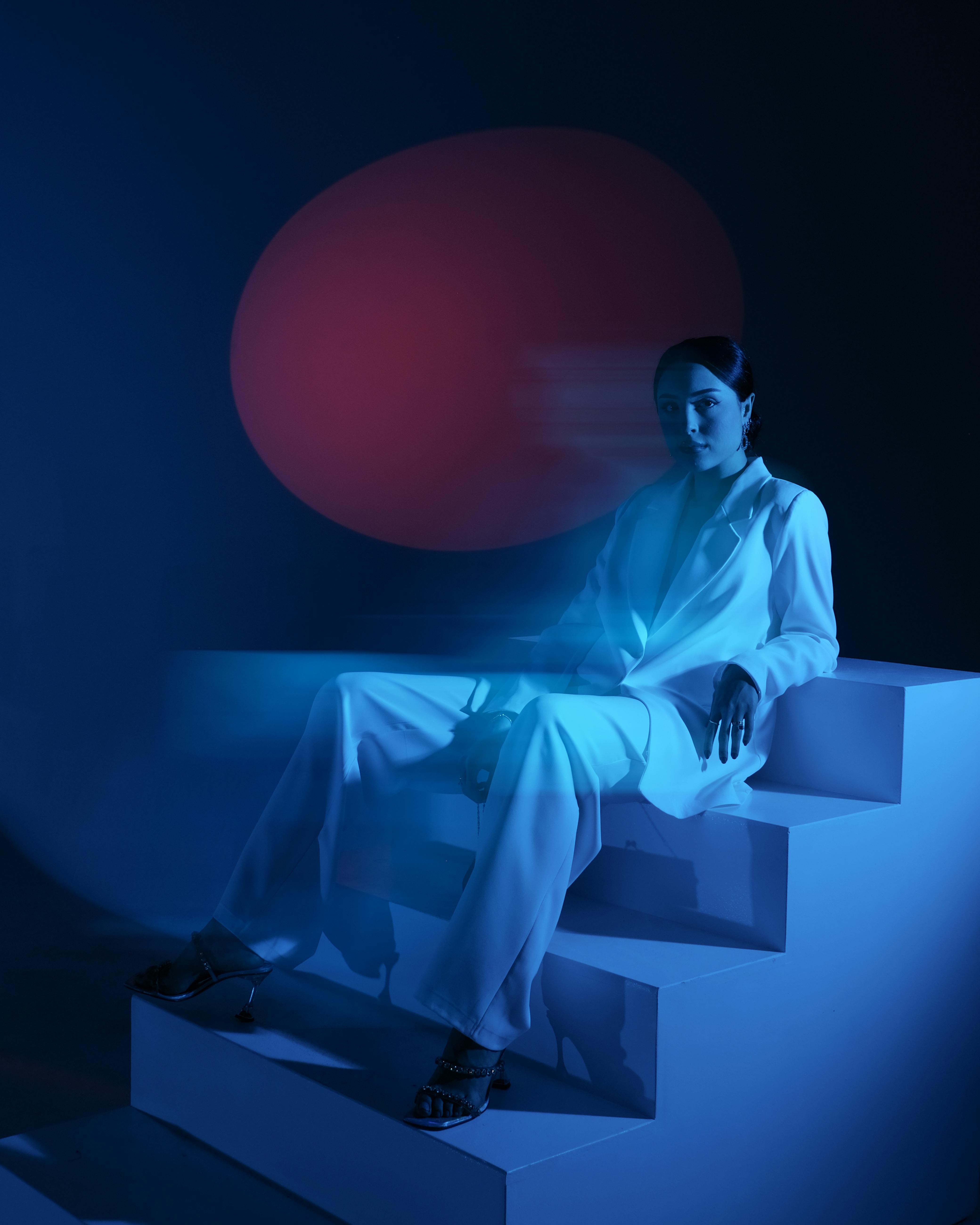 Stylish woman in white sitting on stairs artfully lit with blue light.