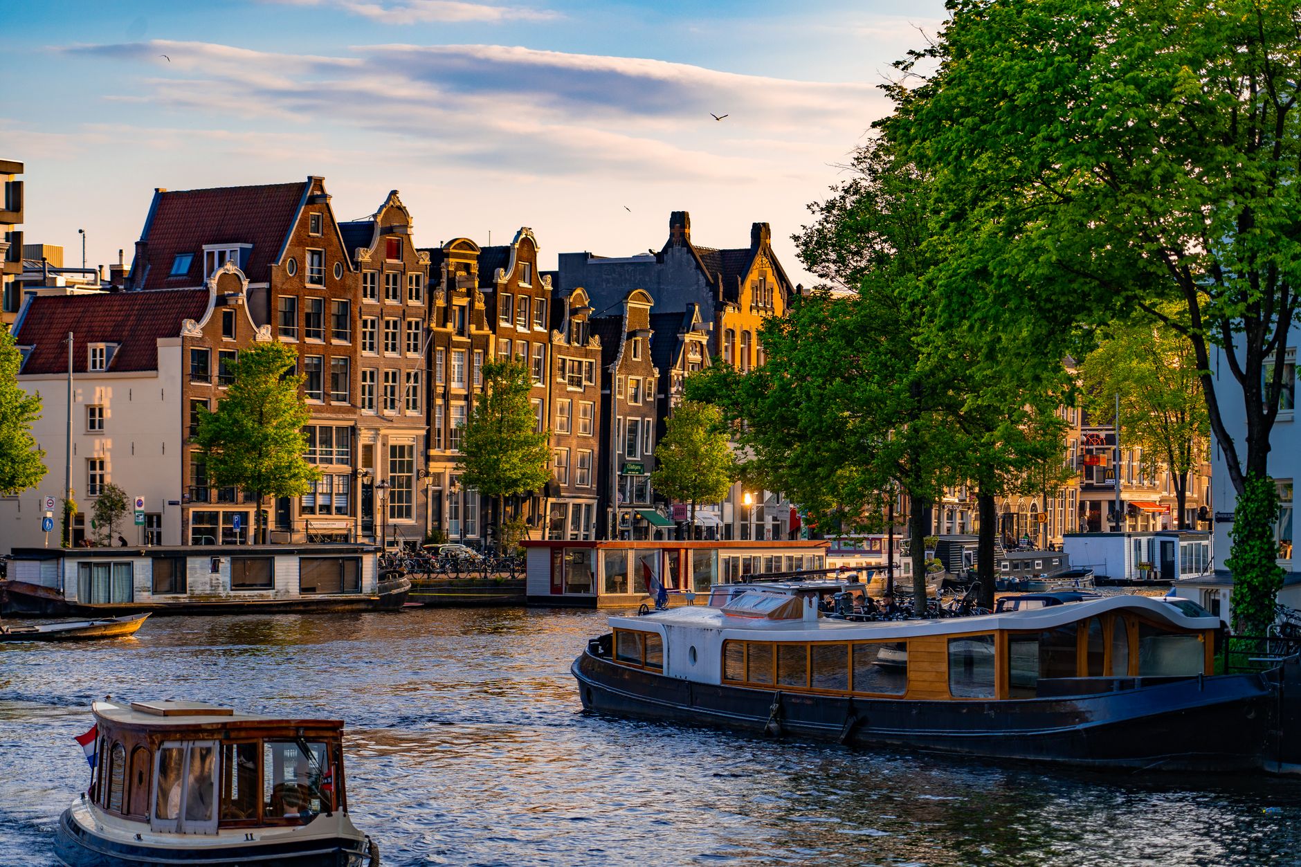 Scenic view of Amsterdam canal houses and boats at sunset with vibrant green trees.