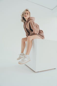 Stylish model in casual hoodie sitting on a white cube against minimal white background.