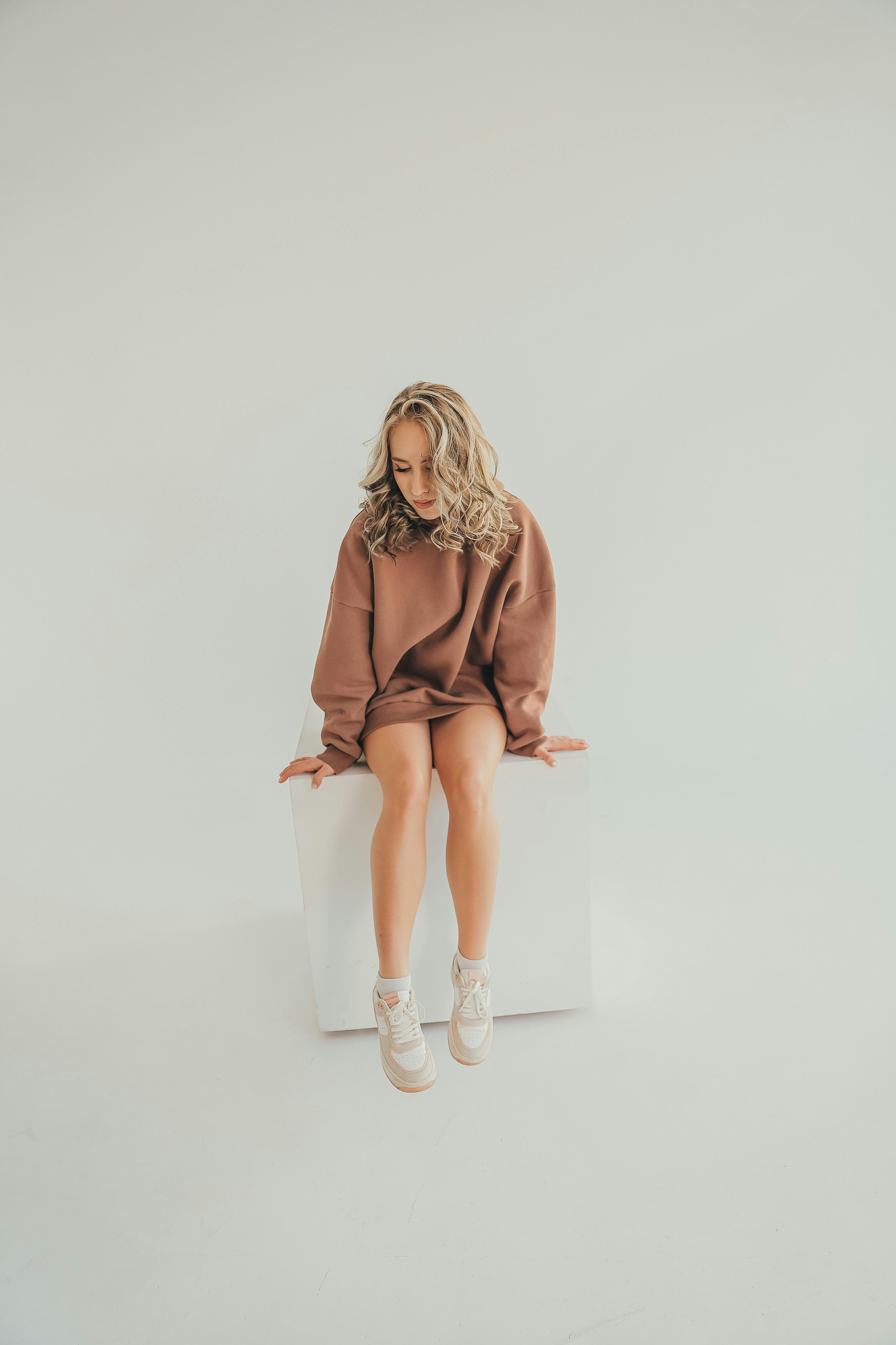 Free A woman in a brown jumper poses on a white cube in a minimalist studio setting. Stock Photo