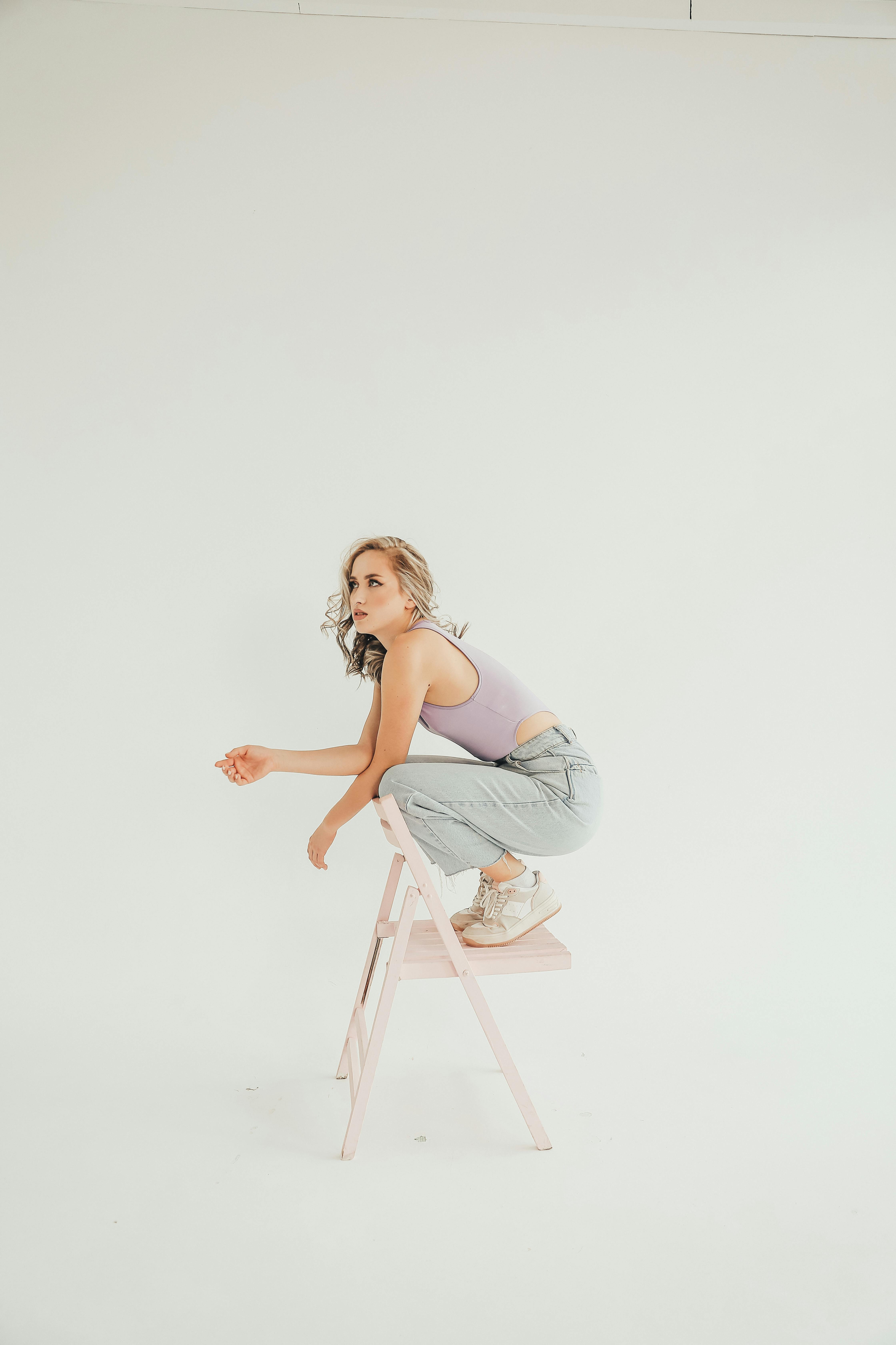 Free Stylish woman in purple bodysuit and jeans posing on stool against white background. Stock Photo
