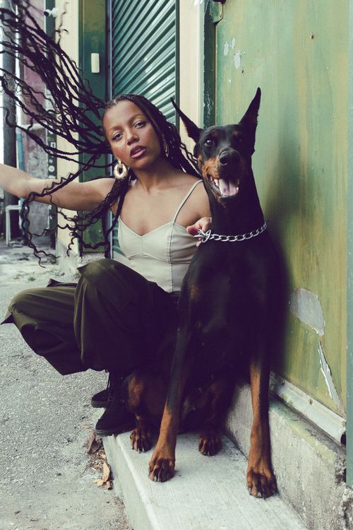 Free A fashionable woman crouches beside a Doberman, showcasing urban street style with braided hair and bold fashion. Stock Photo