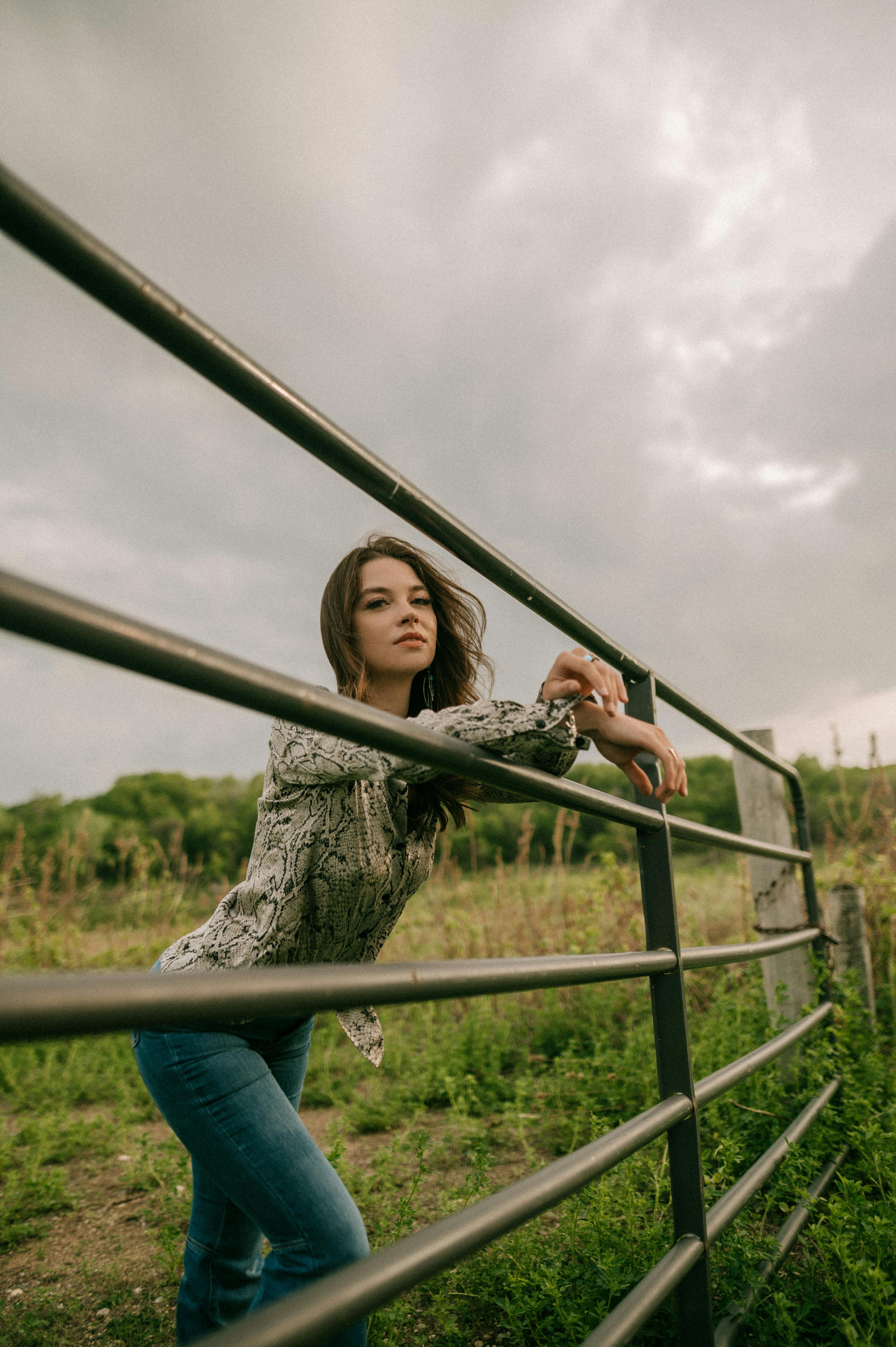 A woman poses thoughtfully leaning on a fence in a lush rural setting under an overcast sky.