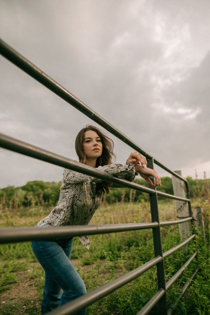 A woman poses thoughtfully leaning on a fence in a lush rural setting under an overcast sky.