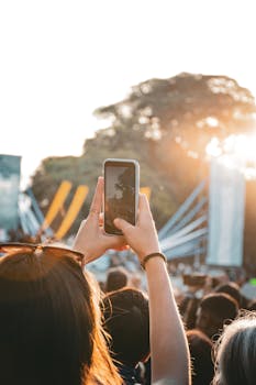 A lively outdoor concert scene with a woman recording the performance using her smartphone under the sunset light.