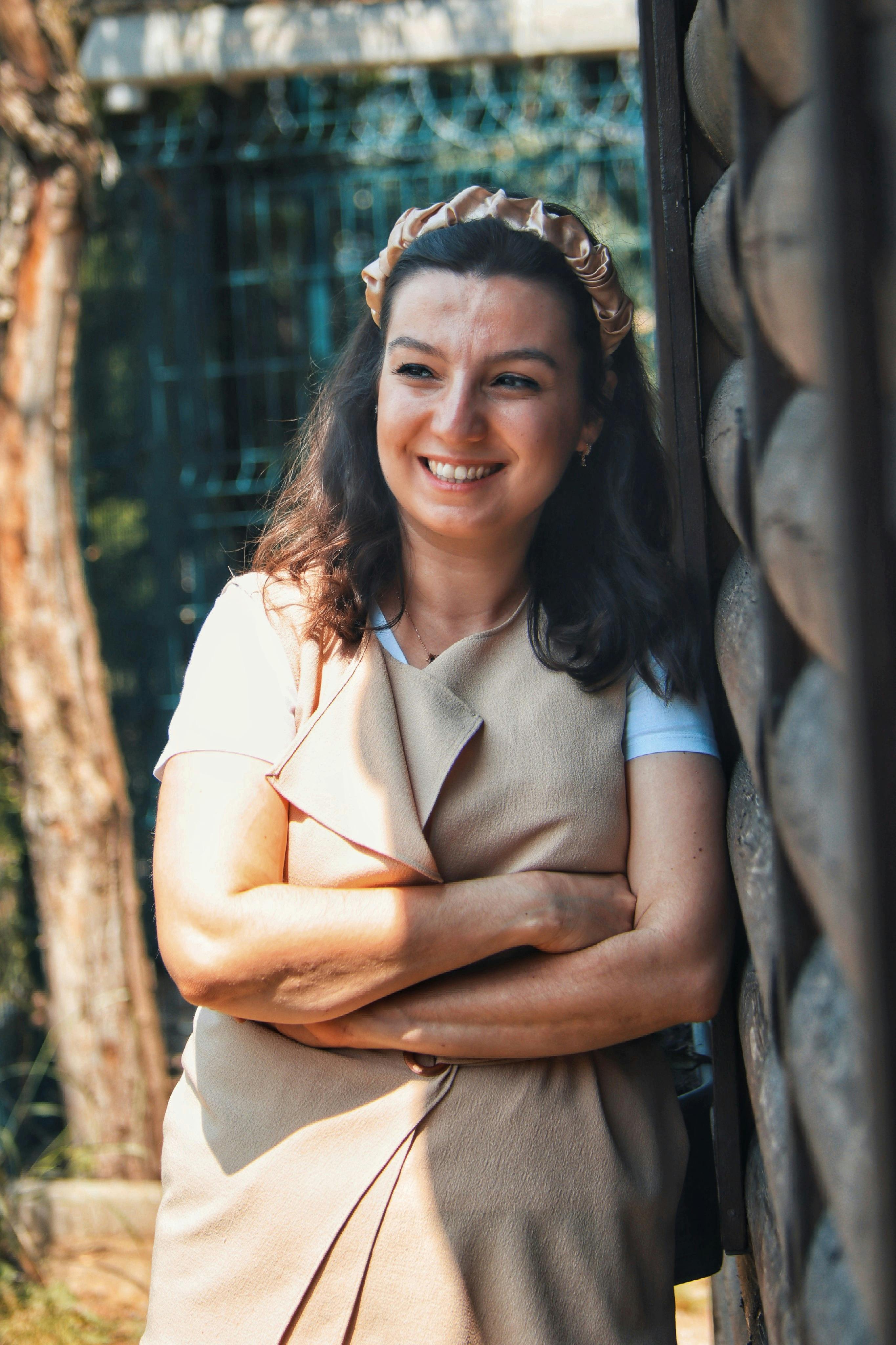 A woman standing and smiling outdoors in warm sunlight, wearing casual fashion with a headband.