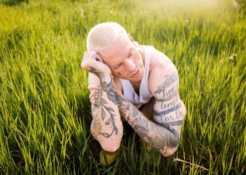 Blonde male crouching in green grass, showcasing tattoos with a serene expression.