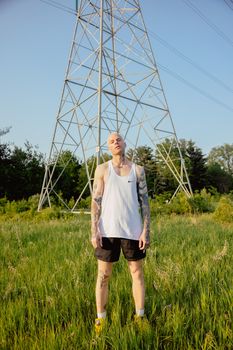 A tattooed man in a tank top stands in a grassy field under power lines on a sunny day.