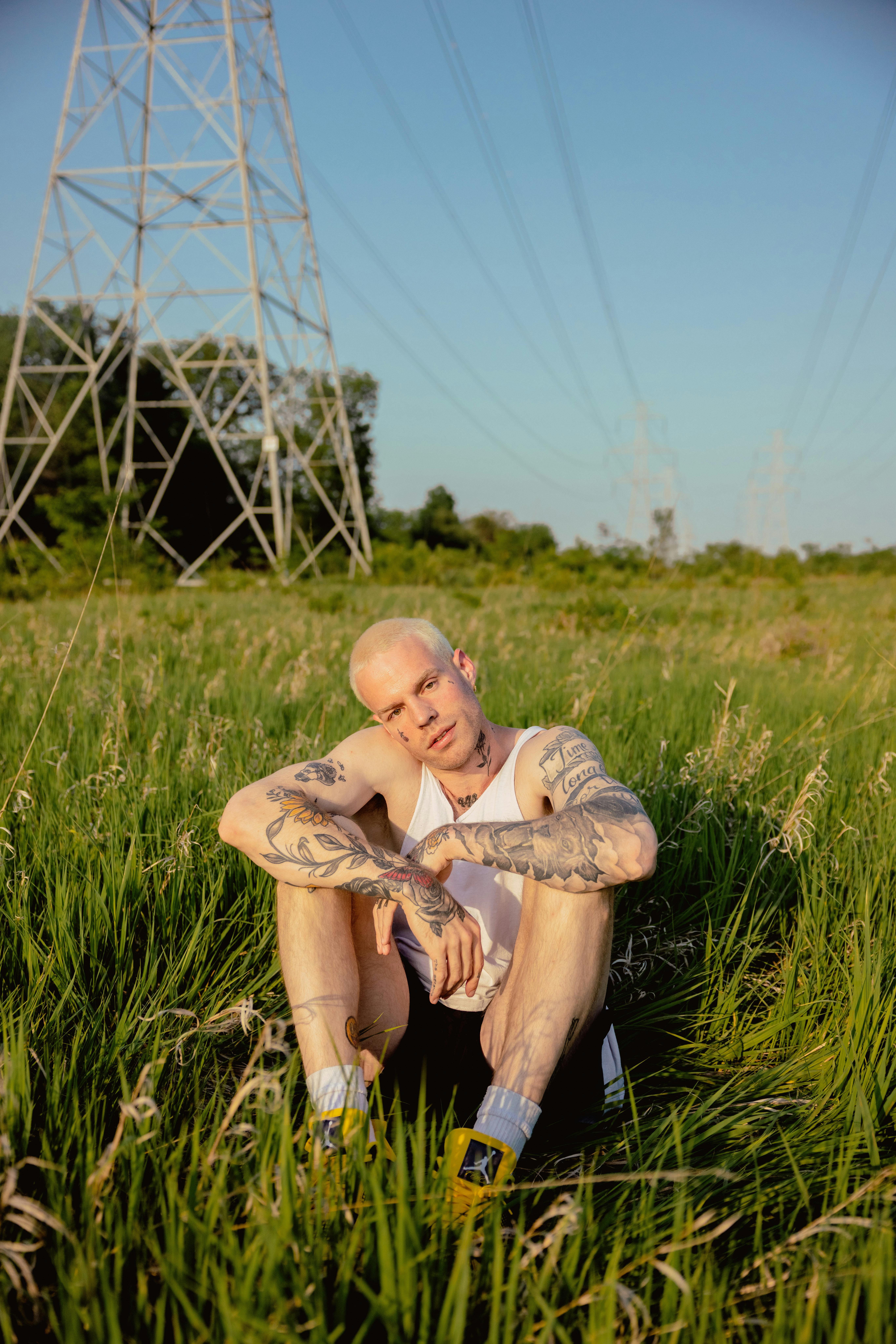 A young tattooed man relaxes in a grassy field under blue skies, enjoying a serene moment.