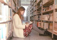 一位亚洲女性坐在图书馆的过道上看书，周围都是书架