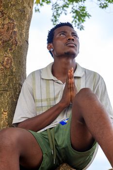 A young man sitting outdoors, clasping hands in prayer beside a tree.