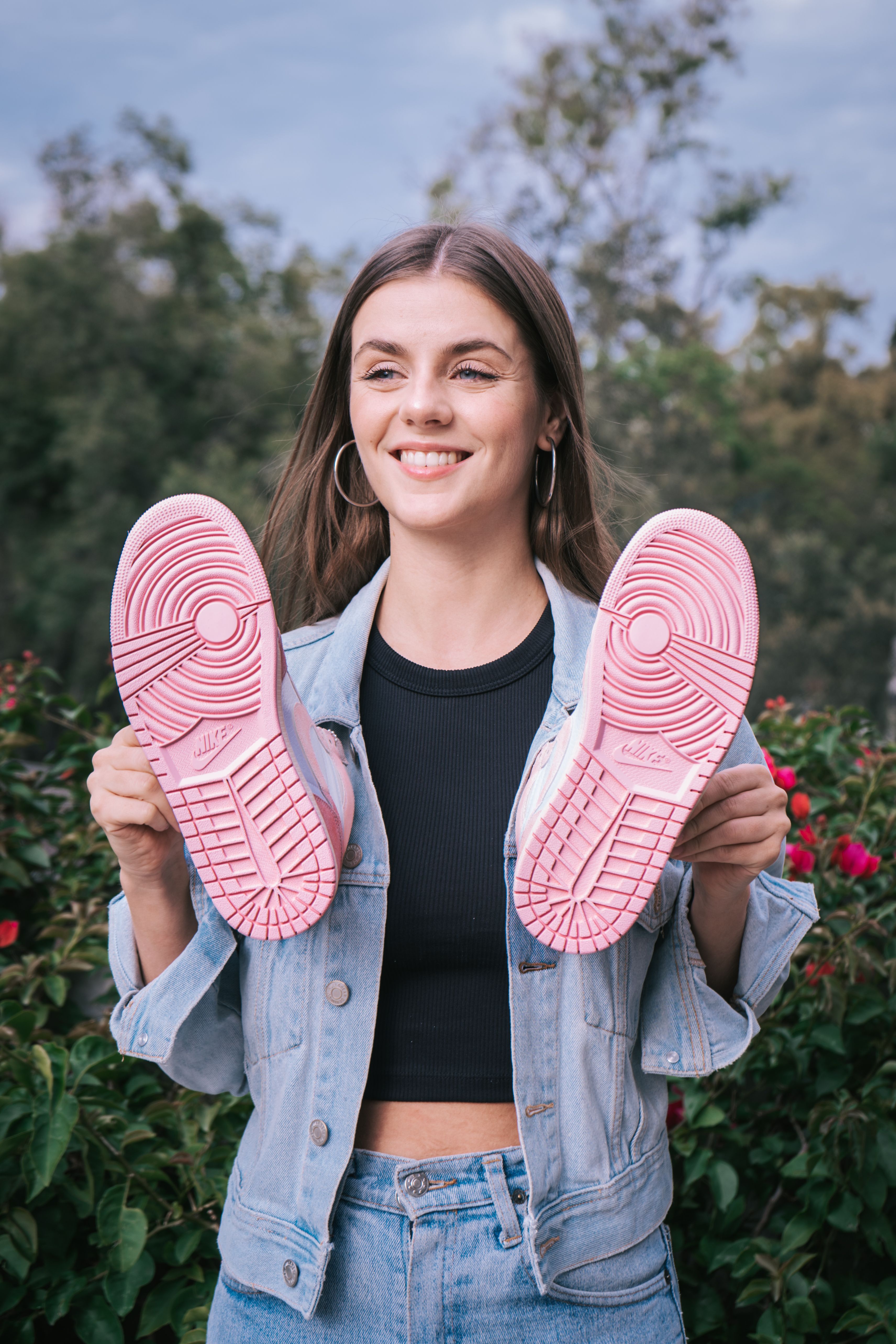 A cheerful woman outdoors holding sneakers with pink soles, showcasing fashion style.