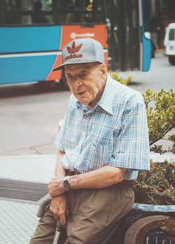 A senior adult man with a cane sits on an urban sidewalk bench in Mendoza, Argentina.