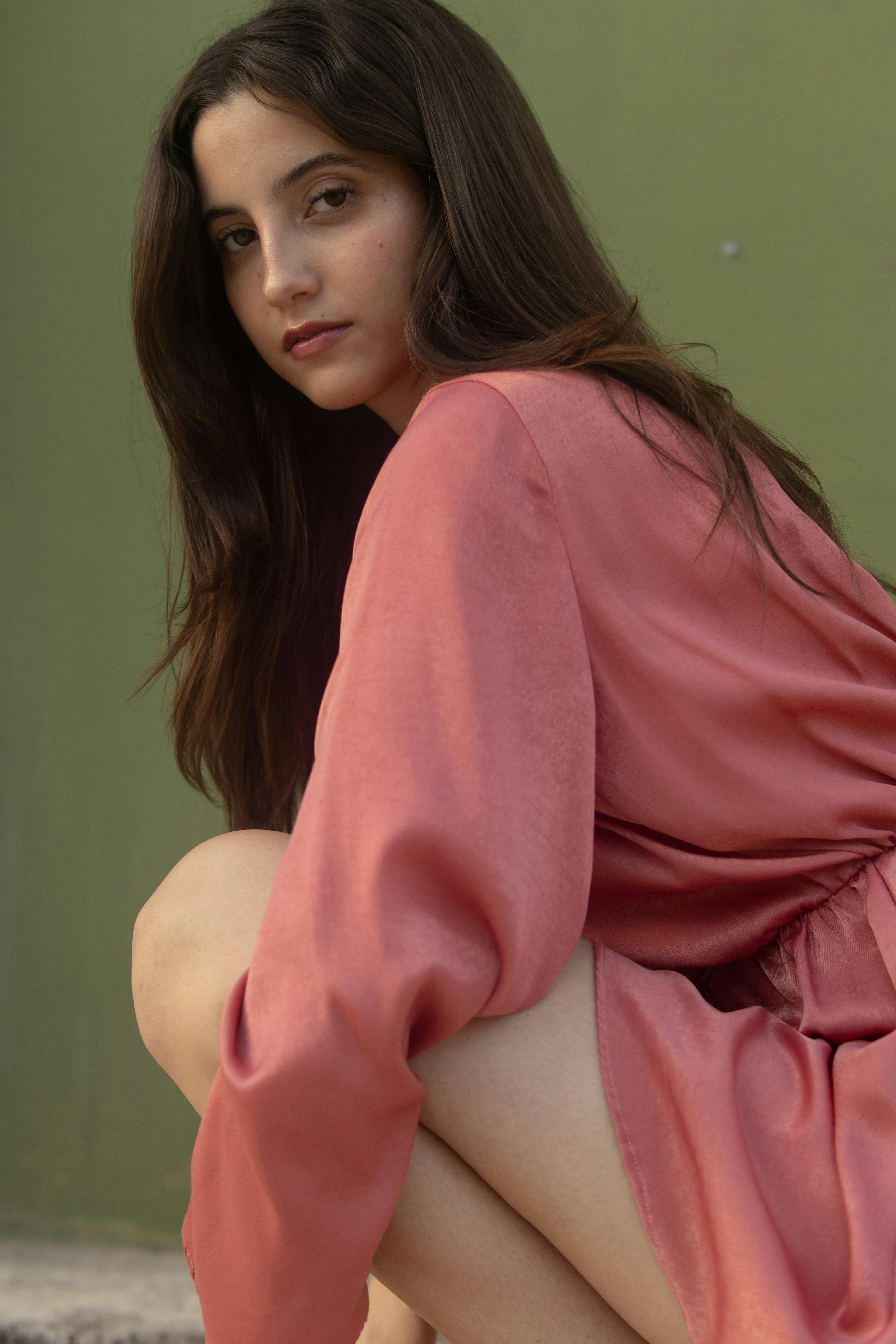 Portrait of a young woman crouching in a pink dress against a green background.