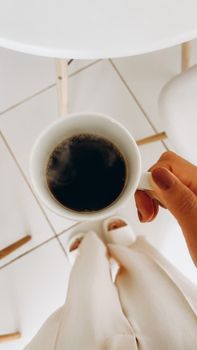 A person holding a steaming cup of black coffee in a modern indoor setting, viewed from above.