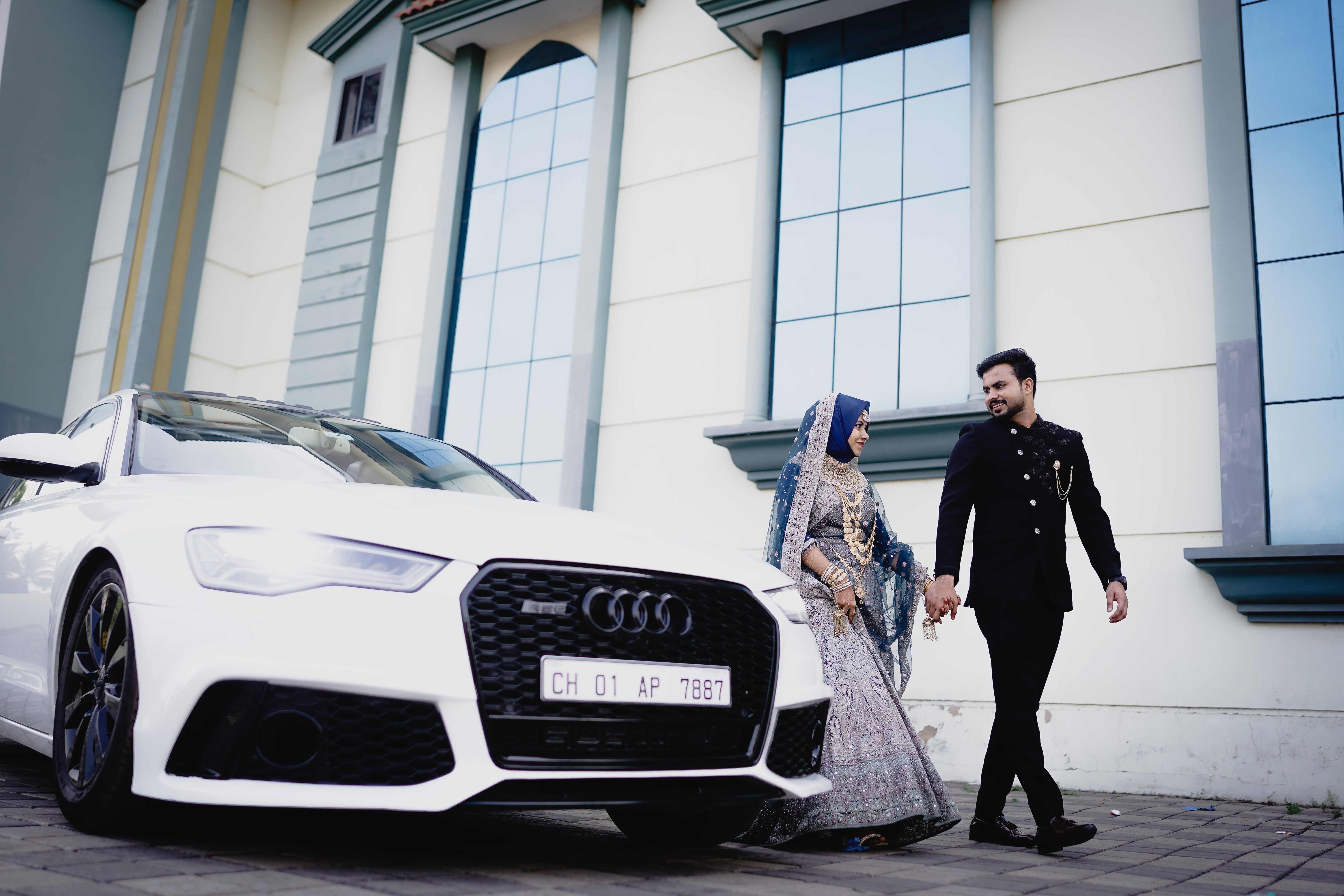 A newlywed couple in traditional attire walks beside a white sports car, showcasing elegance and style.