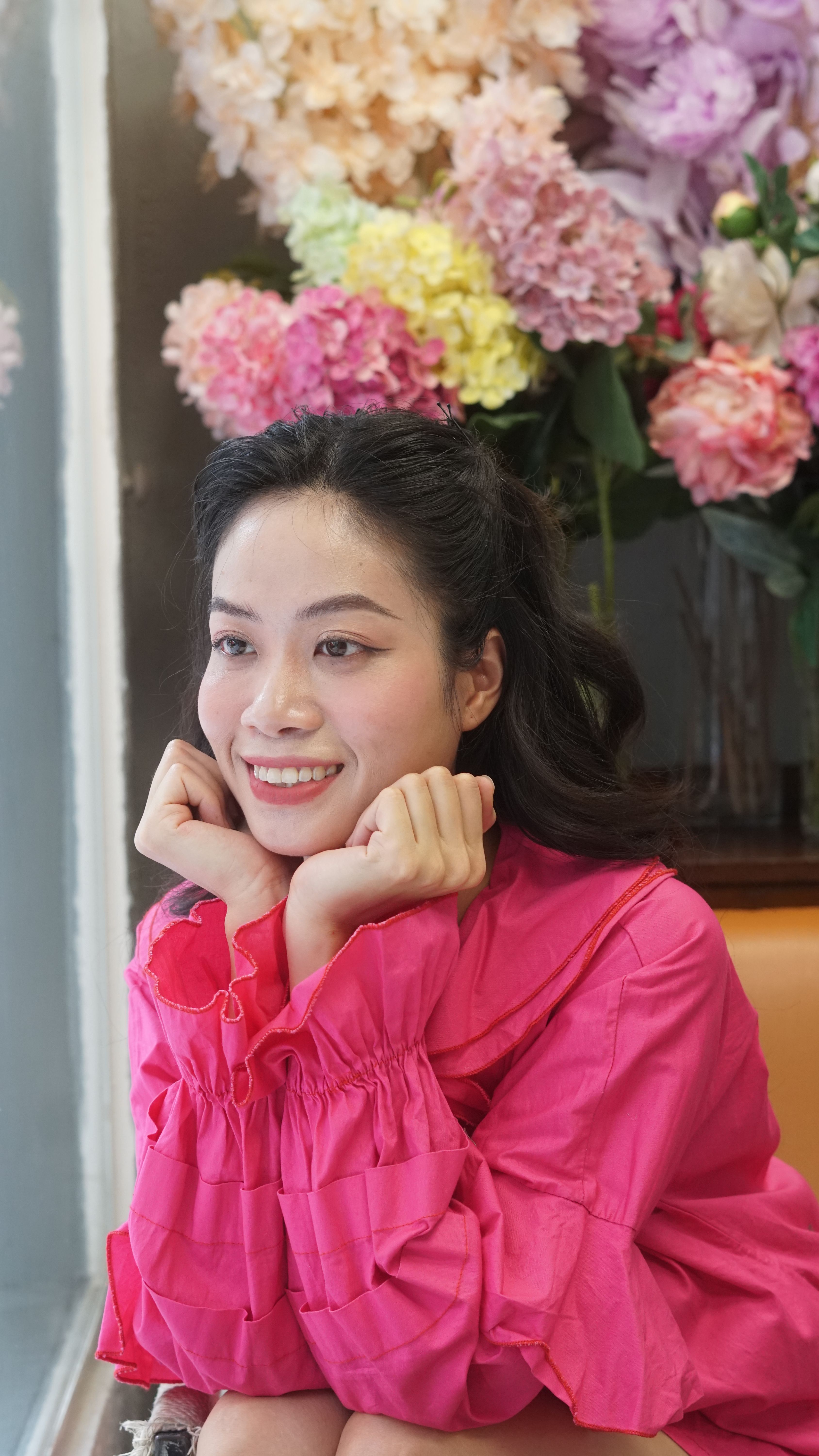 Portrait of a young woman in a pink shirt smiling next to colorful flowers indoors.