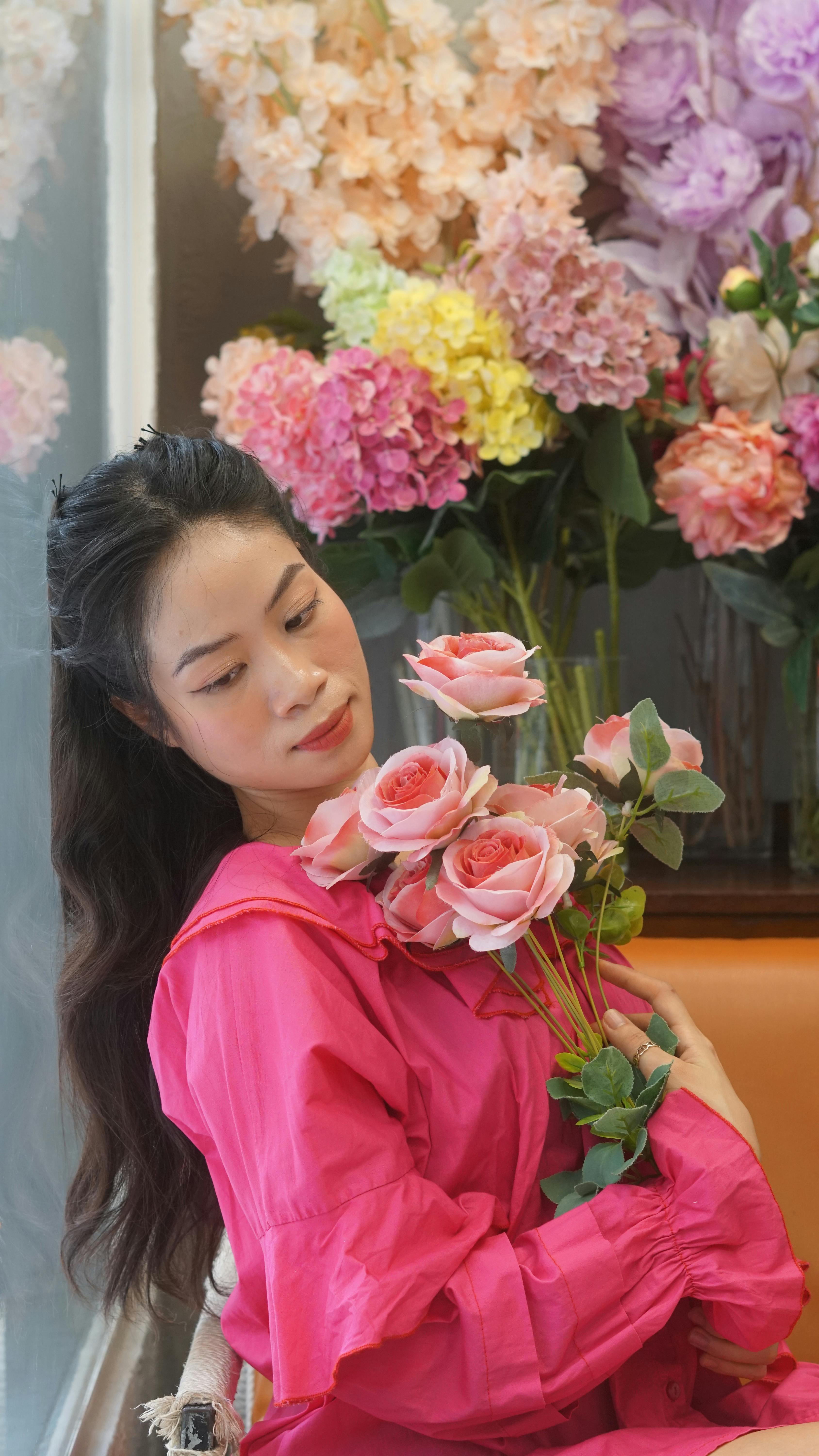 Elegant woman in pink dress holding roses surrounded by vibrant flowers indoors.