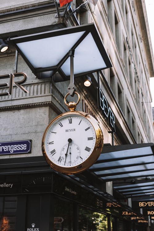 Free A vintage Beyer clock hangs on a bustling city street corner, adding timeless charm to the urban environment. Stock Photo
