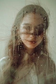 A mesmerizing portrait of a young woman seen through rain-specked glass, creating a dreamy and mysterious effect.