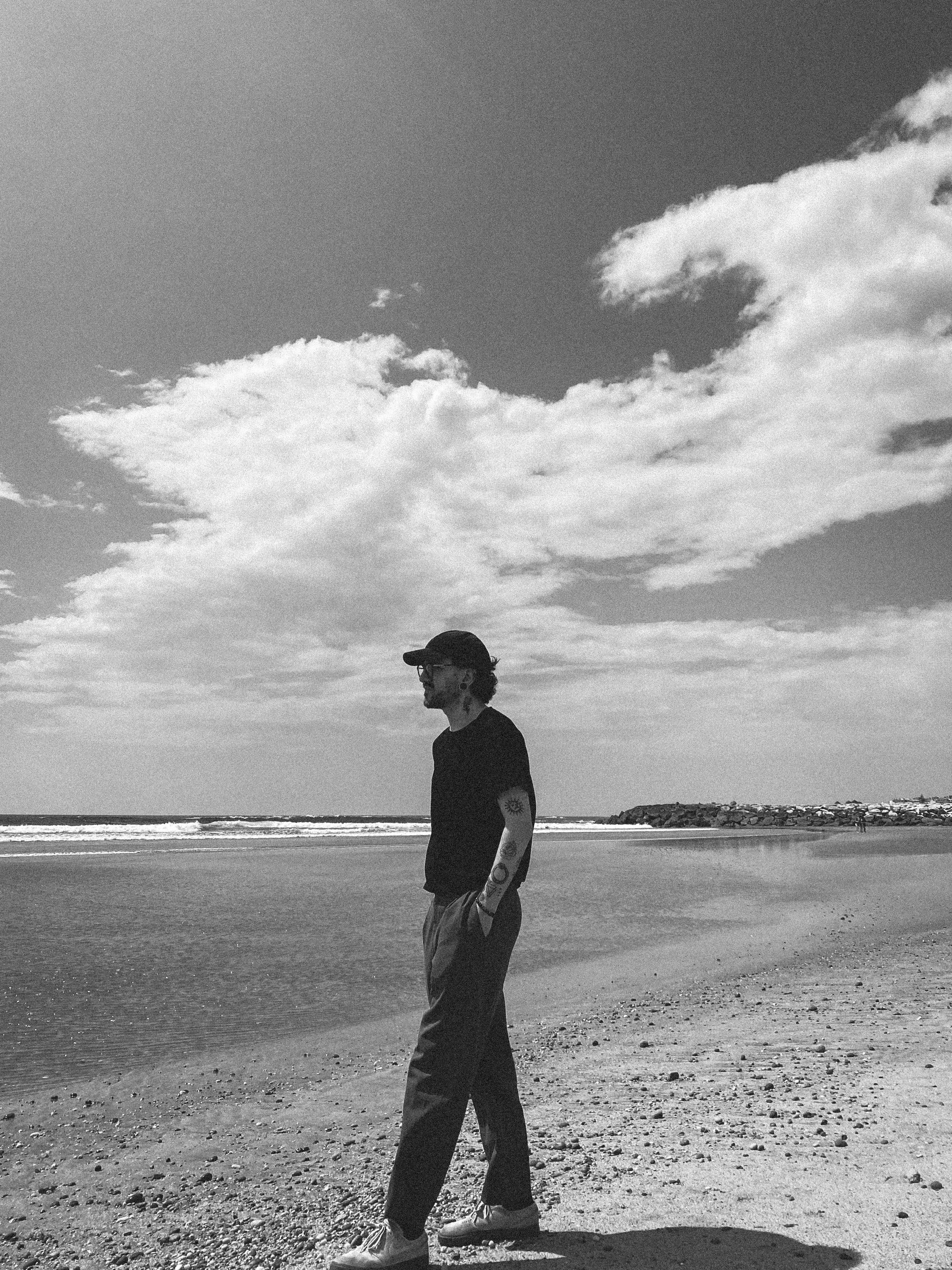 A man in a cap walks along a sunlit beach with clouds overhead, reflecting solitude.