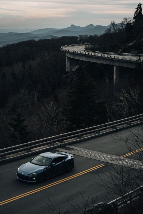 Free A sleek car navigates a winding mountain highway, surrounded by majestic peaks at dusk. Stock Photo